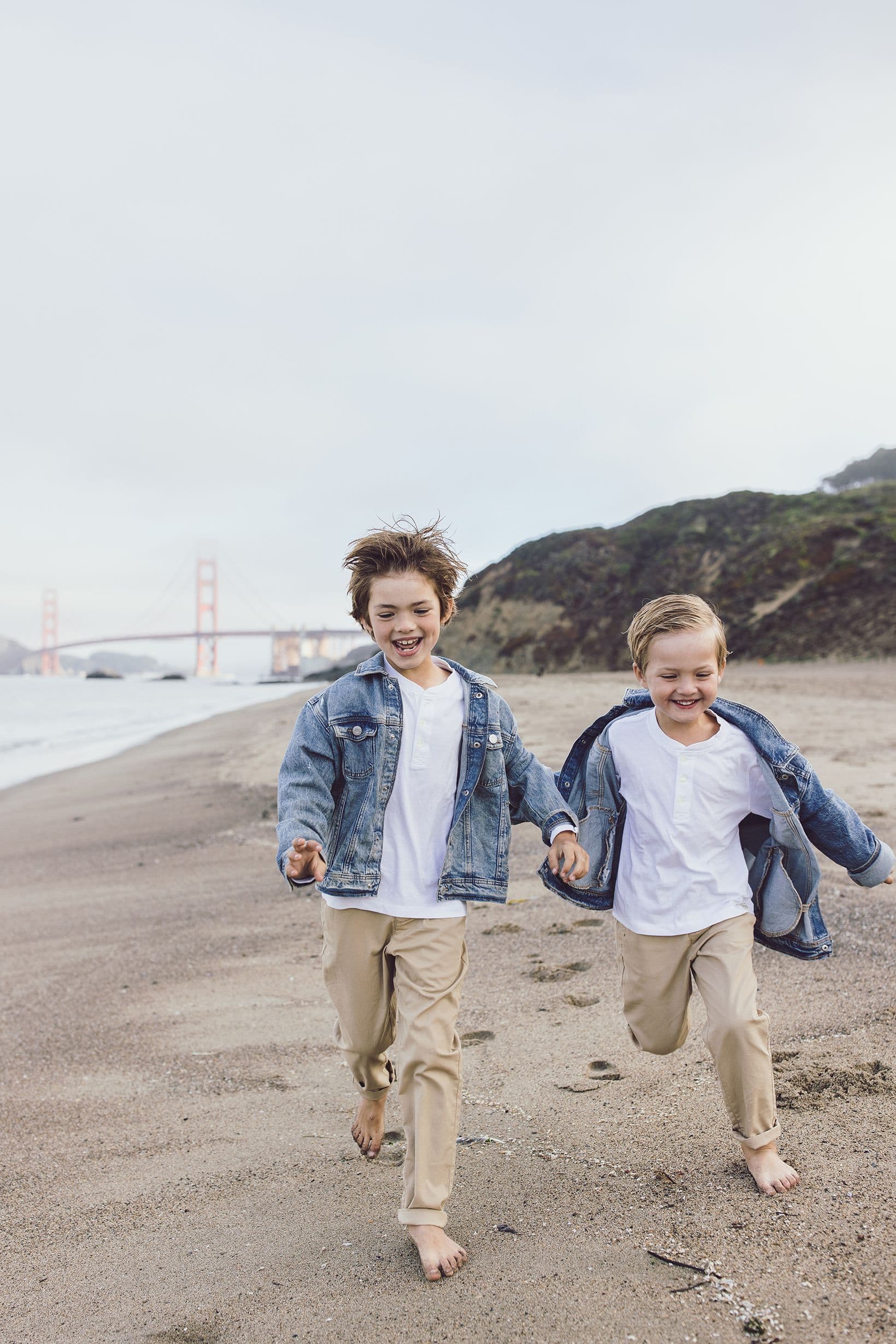 Two boys in denim jackets running barefoot along Baker Beach with the Golden Gate Bridge behind them