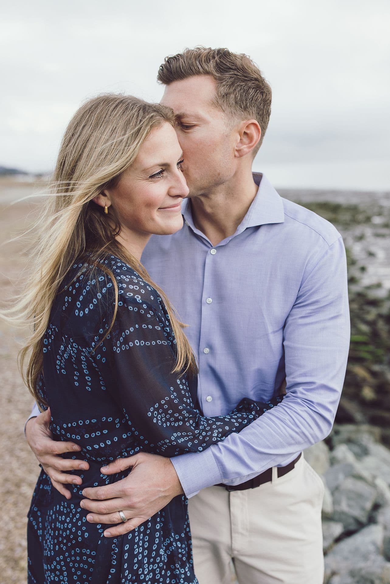 Man kissing his partner's forehead on a pebble beach, her hair lifting in the wind